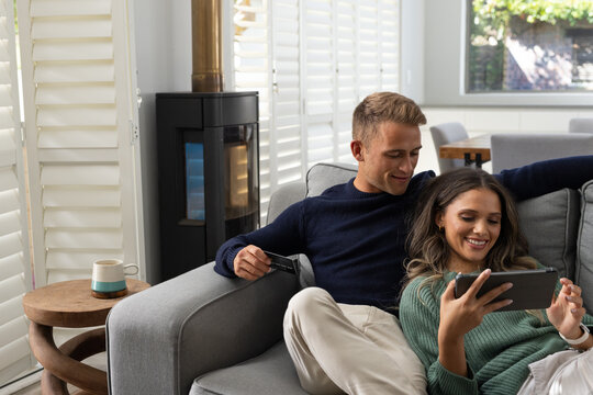 Diverse couple sitting on grey sofa at home, woman reclining holding tablet, man holding card