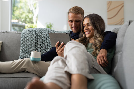 Diverse couple sitting on grey sofa wearing sweaters, leaning and holding tablet with travel mug