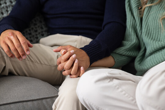 Diverse couple sitting on gray couch, holding hands, wearing knit sweaters and beige pants