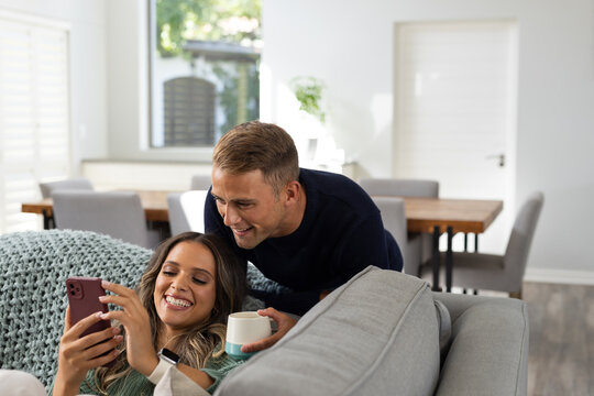 Diverse couple reclining on gray sofa at home, woman smiling at red smartphone, man holding mug