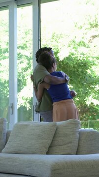 Vertical video: Entering woman greeting man, turning, hugging by glass door with couch, olive shirt