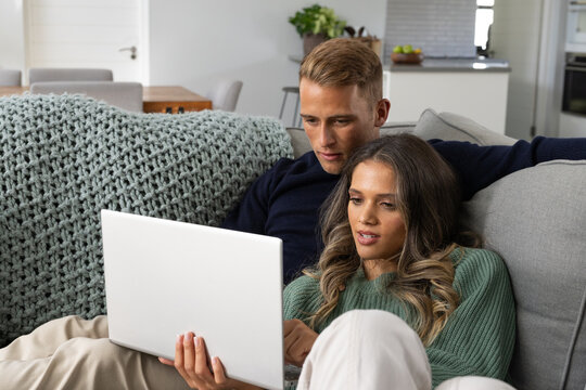 Diverse couple sitting on gray sofa leaning in watching silver laptop at home, chunky throw