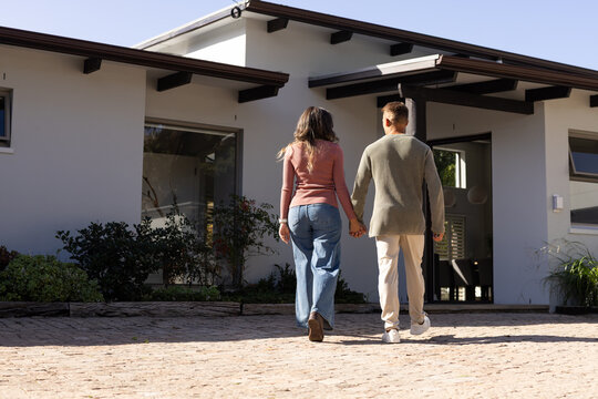 Couple man and African American woman walking hand-in-hand toward modern house on paved driveway