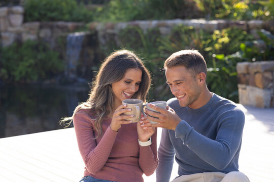 Diverse couple sitting on deck by pond, toasting mugs, mauve top, blue sweater and wristwatch seen