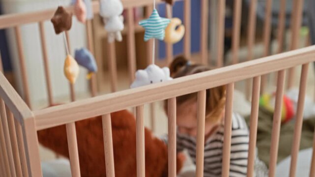 Girl sitting in a crib holding a plush monkey with her hand and gripping the rail inside a building; curiosity play.
