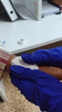 A close up of a healthcare worker wearing blue nitrile gloves applying gauze to a woman's hand in a clinical setting, representing medical care, wound treatment, and patient support during a healthcar