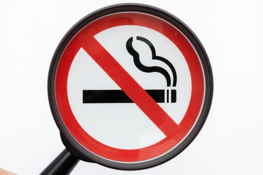Magnifying glass examines no smoking sign, highlighting health risks and public health awareness. Focus on preventing tobacco addiction and promoting smoke-free environments.