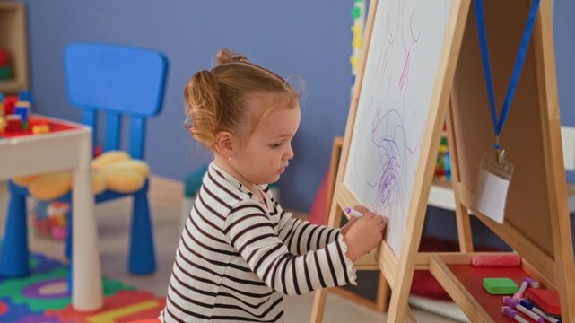 Little blonde girl draws purple scribbles on easel with marker in colorful preschool studio; concentration.