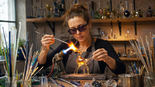 Woman glassblower shaping glass deer figure with torch in workshop