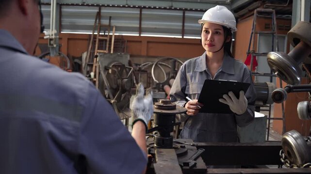 Asian female inspector with a clipboard takes notes while supervising a male coworker repairing machinery components inside a manufacturing factory