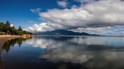 Fototapeta premium Wide angle view of coastal landscape with calm water reflecting trees and mountains under dramatic cloudy sky. Sandy beach borders still lake during daytime.