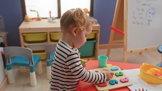 Little blonde girl in striped shirt placing colorful wooden number puzzle pieces with hand on preschool table near easel and toy cups inside a building; quiet concentration.