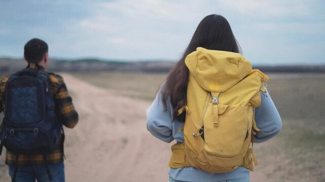 Two backpackers walking on desert road during hiking adventure. Young couple with backpacks hiking on dirt path. Man and woman trekking together. Friends exploring desert trail outdoors.