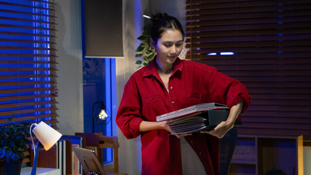 An Asian architect carries a large stack of design files and binders in her office at night.