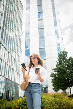 Mature professional woman using smartphone and holding coffee near office towers