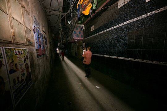 Dhaka, Bangladesh - 03 June 2026: View of a man carrying a child through a narrow dark alleyway with tangled overhead wires and posters on the walls.