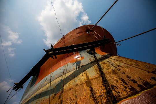 Dhaka, Bangladesh - 03 June 2026: View of a massive ship hull in a shipyard with a worker's shadow cast on the side, featuring heavy chains and scaffolding under a bright blue sky.