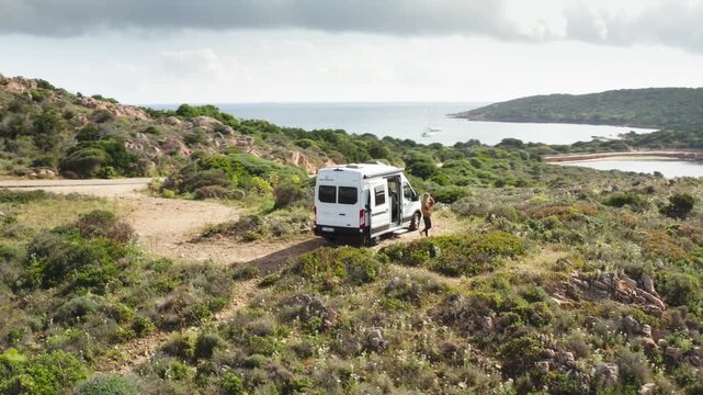 Wide Drone shot of girl walking out of Camper van on mediterranean island as drone circles van overhead