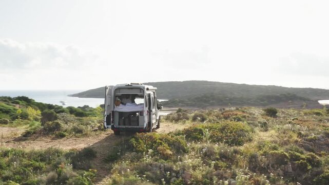 Drone shot of Camper van on mediterranean island with trunk doors open and girl on bed drinking tea and coffee as drone circles van 