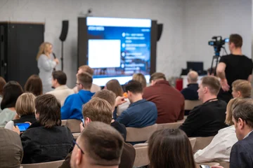 Business conference audience watching presenter on large screen, attendees using phones and camera, candid learning and networking atmosphere. © Anton Gvozdikov