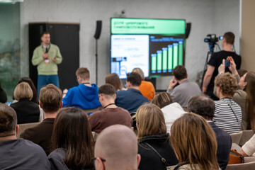 Fototapeta premium Conference audience watches speaker and slide deck, business presentation in modern meeting space, people taking notes and recording on camera.