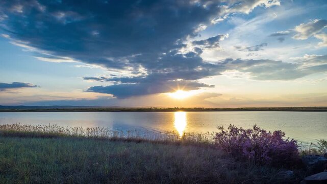 Clouds run across the sky with a sunset light over a lake with herbs on a shore