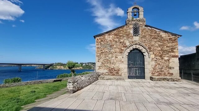A low-angle view of the rustic stone facade of the Chapel of the Most Holy Trinity in Ribadeo, featuring a small bell gable under a deep blue sky with wispy white clouds.