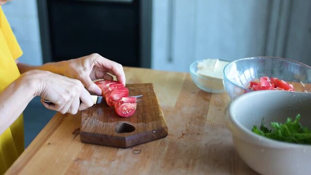Woman chopping tomato on wooden cutting board near fresh herbs in modern video Full HD kitchen