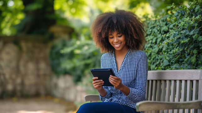 A smiling Black woman with voluminous curly hair sits on a wooden park bench, reading from an e-reader in a black folio case. Lush green trees and stone walls create a peaceful out