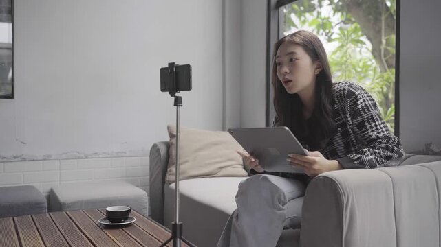 A young woman sits on the right side of the frame on a sofa, speaking toward a smartphone on a stand while holding a tablet