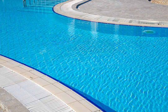 Curved edge of an outdoor swimming pool with clear blue water, white drain grate and stone paving in bright sunlight