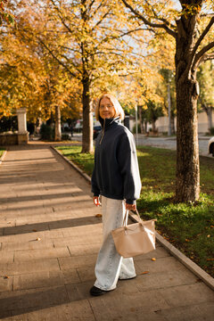 Stylish smiling senior woman 55-60 year old wearing blue sweatshirt and denim pants holding tote bag over sunny autumn background in park. 60s.