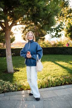 Stylish smiling senior woman 55-60 year old wearing blue sweatshirt and denim pants holding tote bag over sunny autumn background in park. 60s.