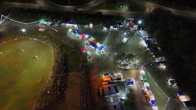 Vista a&eacute;rea de fiesta gaucha nocturna con p&uacute;blico y pista ecuestre, carrera de barriles en el campo argentino