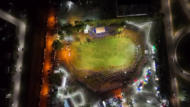 Vista a&eacute;rea de fiesta gaucha nocturna con p&uacute;blico y pista ecuestre, carrera de barriles en el campo argentino