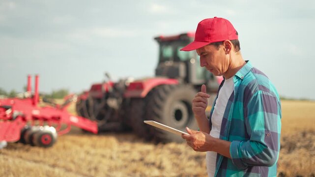 Modern farmer in red cap and plaid shirt uses digital tablet to manage agricultural operations in cultivated field. Large tractor with red cultivator equipment visible in background farmland.