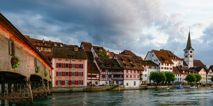 Historic buildings line the riverside with a church in the background and clouds in the sky during the evening