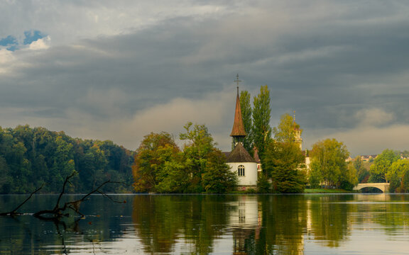 A view of a small church surrounded by trees and water under a cloudy sky in a serene setting