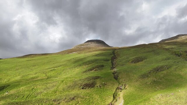 A dramatic aerial drone shot pushing forward and booming up towards the Pap of Glenco mountain in the Scottish Highlands, UK. The slopes are vibrant green under heavy clouds on a spring afternoon.