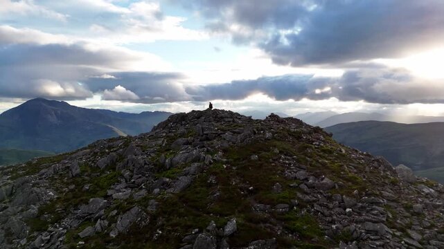 A drone executes a push-in, boom up motion, closing in on a solitary figure on the rugged summit of Pap of Glencoe in the Scottish Highlands.