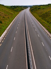 Obraz premium Long, empty highway stretches through green countryside. Road curves gently under cloudy sky. Grass and shrubs line both sides of the road. Distant hills rise behind the road. Travel and tourism,