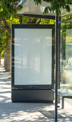 Blank advertising billboard mockup integrated into a modern bus shelter on a tree-lined street in Lisbon, Portugal. The empty vertical display panel provides ample copy space for branding, promotional © Pedro