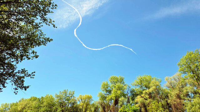 White aircraft contrail creating a curved pattern in a clear blue sky above green spring trees.