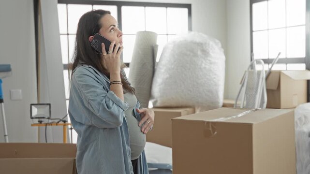 Pregnant woman holding smartphone to ear and cradling belly in a building surrounded by moving boxes and packing materials; hopeful anticipation.