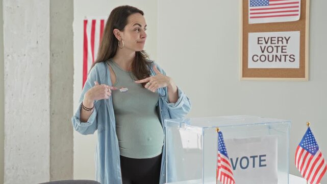 Pregnant woman smiling points to an i voted sticker on her chest at a polling building beside a transparent ballot box and flags; civic duty pride.
