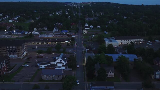 Cinematic aerial drone shot following above a main street in Truro, Nova Scotia, Canada at dusk. Footage features moving cars, residential homes, illuminated streets, and evening Maritime city