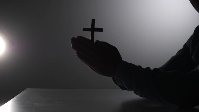 Silhouette of a person with hands clasped in prayer, holding a wooden cross, symbolizing faith, hope, spirituality, and religious devotion against a dark background with light