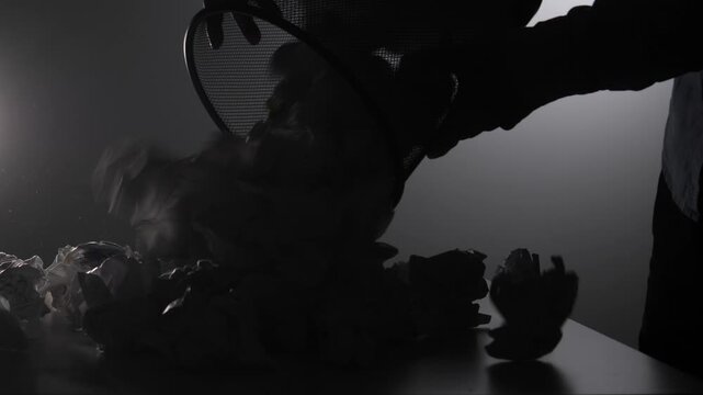 Silhouette of a hand discarding numerous crumpled pieces of paper into a small wire wastebasket, concept of failure, rejection, writer's block, and effort in a creative process