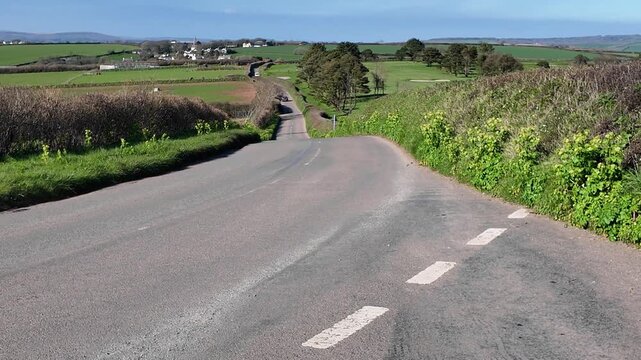 Bigbury south Devon England UK. 27.03.2026. Video. A car travels along a winding asphalt road bordered by vibrant green hedgerows and fields under a clear blue sky