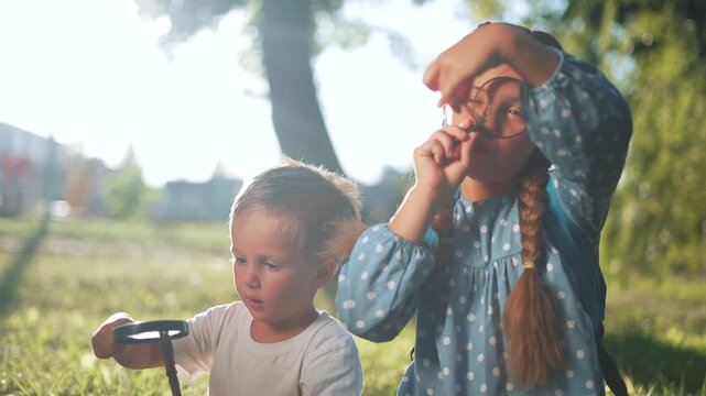 Children explore nature with magnifying glass in sunny park. Girl with braid looks through magnifying glass closely. Boy holds stick and magnifying lens on grass. Kids exploring outdoors in sunlight.
